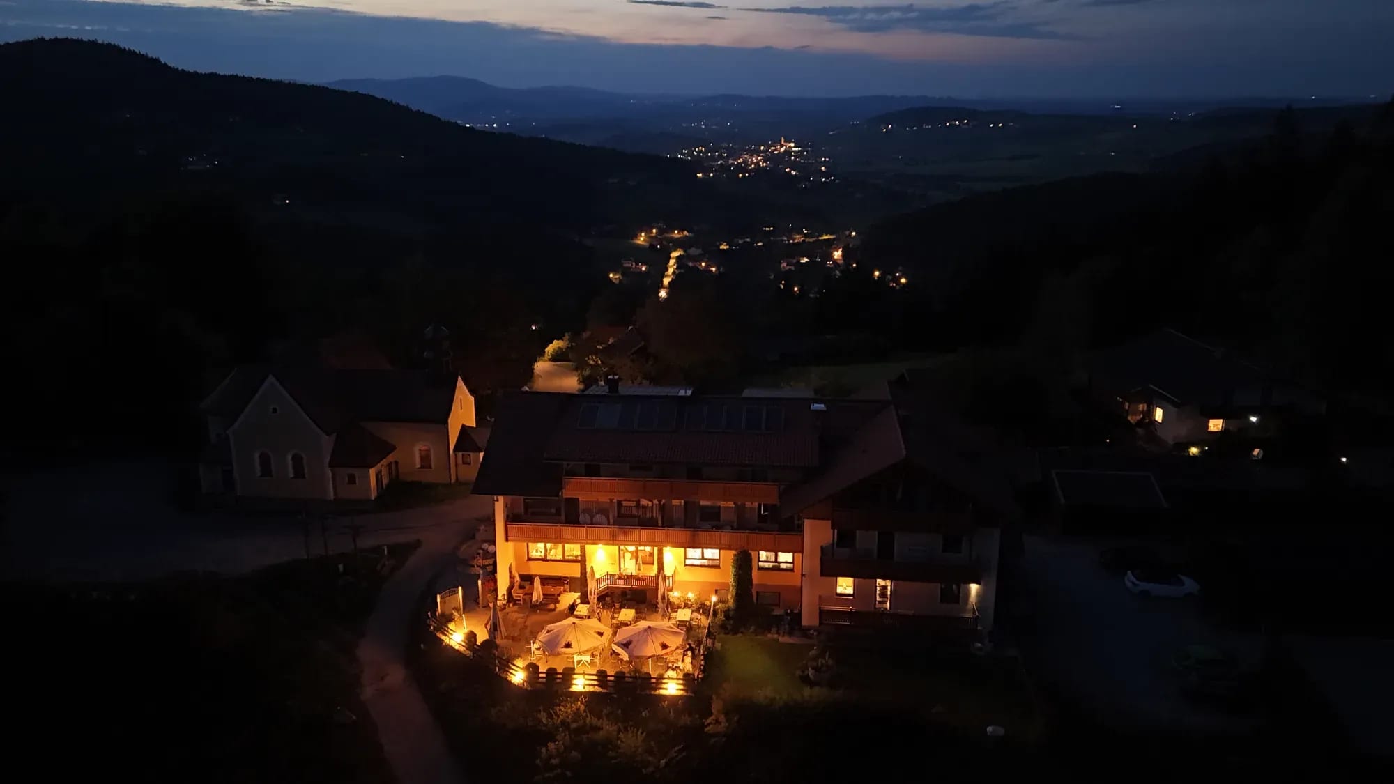 Der Kolmsteiner Hof in der Abenddämmerung — beleuchtete Terrasse, darunter die Lichter des Tals und die Berge des Bayerischen Waldes
