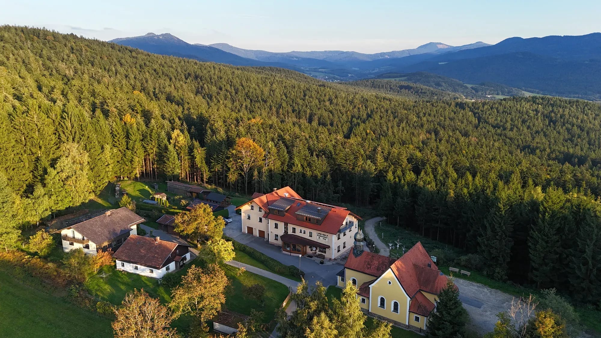 Luftaufnahme des Kolmsteiner Hofs im Abendlicht — umgeben von Wald, dahinter das Bergpanorama des Bayerischen Waldes